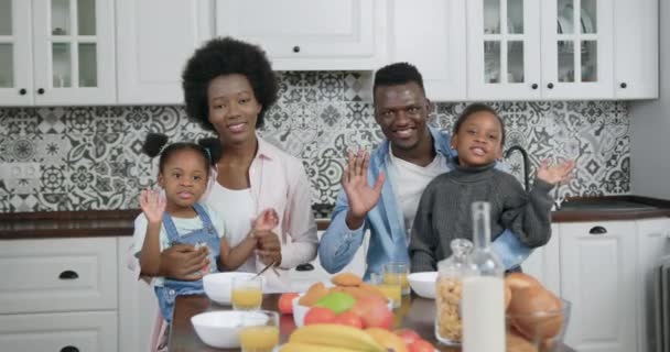 Портрет прекрасної усміхненої щасливої у високому дусі афроамериканської сім'ї, яка дивиться на камеру в сучасній кухні і махає руками під час сніданку, повільний рух — стокове відео