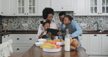 Çekici, pozitif gülümseyen, mutlu, Afro-Amerikan ailesi yemek masasında oturup tablet bilgisayar kullanırken küçük kızlar kurabiye yiyor.
