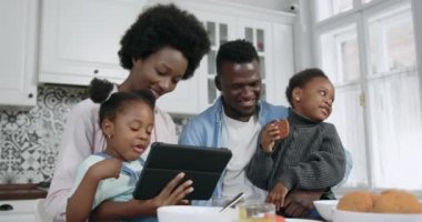 Çağdaş mutfaklarda yemek masasında otururken tablet kullanan küçük kızları olan mutlu, güler yüzlü, Afro-Amerikan çifti, kapayın, 4k