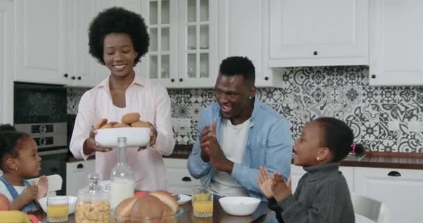 Hermosa feliz sonriente familia de piel negra disfrutando del desayuno conjunto en la cocina contemporánea y regocijándose sabrosas galletas que la madre pone en la mesa para el té, de cerca, 4k — Vídeos de Stock