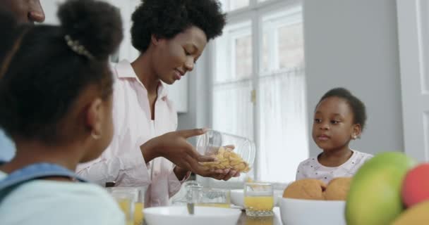 Linda satisfeita sorrindo jovem mulher de pele preta derramando flocos de milho em tigela, preparando café da manhã para suas filhas pequenas bonitos que se sentam em torno da mesa da cozinha, 4k — Vídeo de Stock