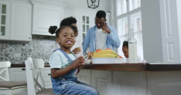 Lovely smiling satisfied small african american girl in jeans overalls sitting near kitchen table on her familys background and looking at camera, 4k — стоковое видео