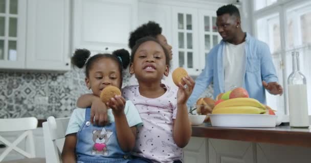 Vonzó mosolygós boldog kis afro-amerikai lányok pózolnak a kamera a kortárs konyhában a háttérben beszélő szülők, elölnézet, lassított felvétel — Stock videók