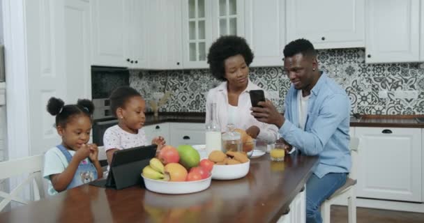 Conceito de família onde belo feliz sorrindo jovem casal de pele preta revisando aplicativos no telefone e suas duas filhas pequenas que usam tablet pc após o café da manhã na cozinha moderna — Vídeo de Stock