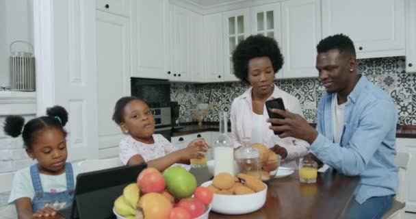 Primer plano de la familia afro-americana amigable sin preocupaciones de buen aspecto que se sientan juntos en la mesa de la cocina y el uso de teléfono inteligente y tableta PC para ver aplicaciones interesantes, cámara lenta — Vídeos de Stock