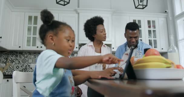 Милі маленькі задоволені чорношкірі дівчата сидять перед планшетним ПК на сучасній кухні, а їхні батьки використовують смартфон поруч з ними, повільний рух — стокове відео