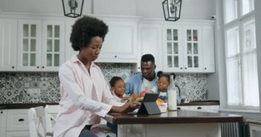 Oldukça mutlu gülümseyen Afro-Amerikan kadın mutfak masasında oturup tablet üzerinde çalışırken yakışıklı kocası küçük kızlarıyla sohbet ediyor, ağır çekimde.