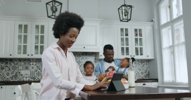 Atractiva de buen humor sonriente madre afroamericana feliz trabajando en la computadora de la tableta en la mesa de la cocina, mientras que sus dos hijas pequeñas lindas sentadas en las rodillas de los padres, de cerca, 4k — Vídeos de Stock