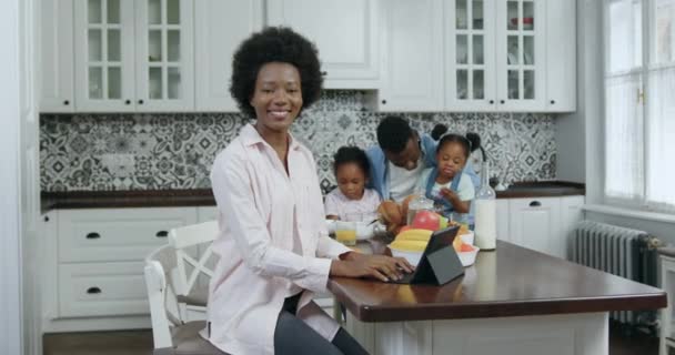 Retrato de hermosa feliz sonriente 30-35aged mujer de piel negra que posa en la cámara en la cocina contemporánea en el fondo de su marido que habla con dos niños lindos, 4k — Vídeos de Stock