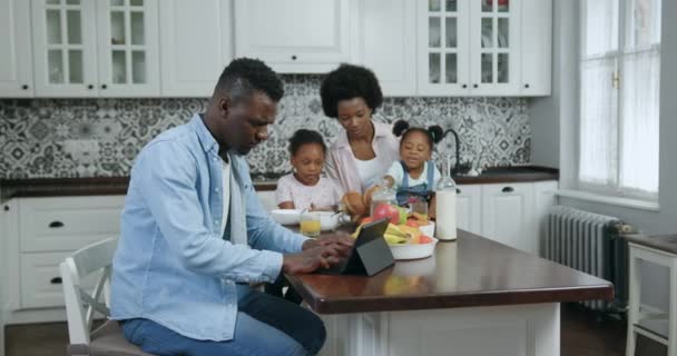 Guapo amante satisfecho hombre de piel negra trabajando en la tableta en la mesa de la cena en la cocina moderna, mientras que su esposa bastante feliz sosteniendo de rodillas a sus dos hijos que desayunan, cámara lenta — Vídeos de Stock