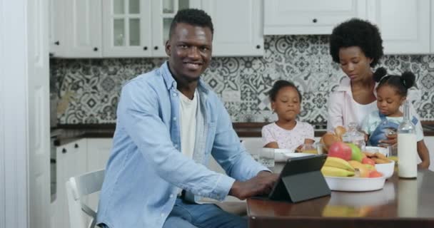 Likable satisfied cheerful 35-aged black-skinned man looking at camera with happy face while his loving wife taking care of their daughters near him in cozy kitchen, close up, 4k — стоковое видео