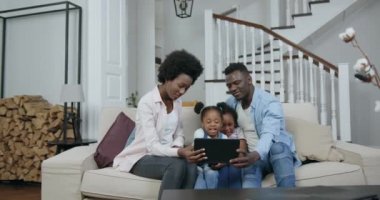 İyi görünümlü, mutlu, küçük kızları olan Afro-Amerikan aileleri oturma odasında rahat bir koltukta oturup çocuk kanalını izlemek için tablet bilgisayar kullanıyorlar.