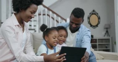 Güzel görünüşlü, mutlu, kaygısız, Afrikalı, küçük çocukları olan, oturma odasında rahat bir koltukta oturan ve ilginç uygulamaları izlemek için tablet kullanan bir aile.