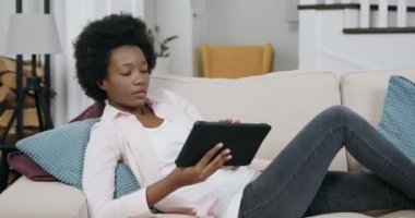 İyi halden oldukça emin, güncel dairedeki yumuşak koltukta uzanan ve tablet bilgisayarını kullanan, ellerini uzatıp çalışmaya devam eden gündelik giysili Afro-Amerikan kadını.