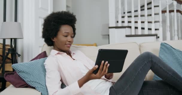 Hermosa sonriente mujer negra moderna feliz con peinado africano acostado en un cómodo sofá en la sala de estar y disfrutando de la revisión de aplicaciones interesantes en el dispositivo de la tableta, vista frontal, 4k — Vídeos de Stock