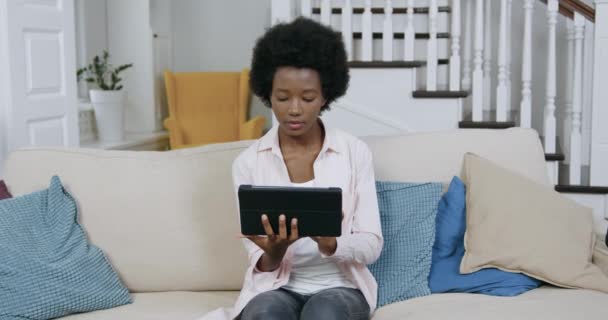 Goed uitziende gelukkig glimlachende jonge moderne zwarte vrouw met Afrikaans kapsel werken op tablet pc, zitten op comfortabele bank en kijken naar de camera, close-up — Stockvideo