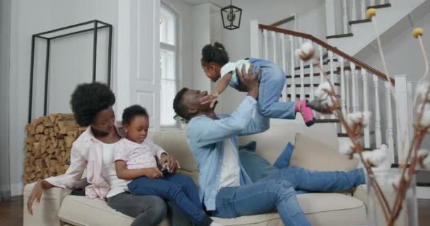 Bonito hombre negro amoroso feliz levantando a la hija en sus manos, sentado en un cómodo sofá en la sala de estar, mientras que su esposa utiliza el móvil junto con otra hija, concepto familiar, 4k — Vídeos de Stock