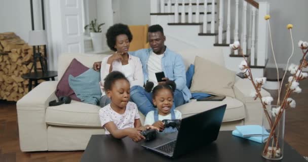 Leuke vrolijke zwarte kleine meisjes spelen videogames op laptop en gebruik gamepads, terwijl hun moeder en vader zitten op een comfortabele bank in de buurt van hen in de woonkamer en browsen telefoon apps, 4k — Stockvideo