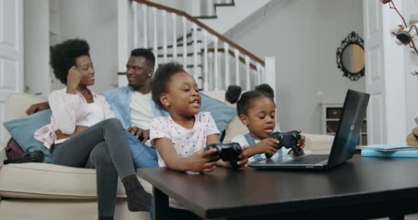 快適な笑顔アフリカ系アメリカ人のカップルリビングルームで柔らかいソファに座って話をしながら、彼らの2人の小さな喜びの娘はジョイスティックでラップトップでゲーム,家族のレジャーコンセプト — ストック動画