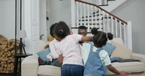 Aranyos boldog mosolygós fekete két lány fut a szüleikhez, akik ülnek kényelmes kanapén a nappaliban és ölelkeznek velük, elölnézet, lassított felvétel — Stock videók