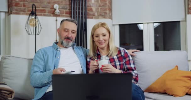 Atractiva sonrisa despreocupada 40-50s pareja relajarse en el cómodo sofá en la casa contemporánea y ver interesante programa en el ordenador portátil — Vídeo de stock