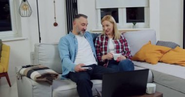 Oturma odasındaki yumuşak kanepede oturup dizüstü bilgisayardan eğlenceli programları izlerken sohbet eden sevimli evli ve neşeli çiftin ön görüntüsü.