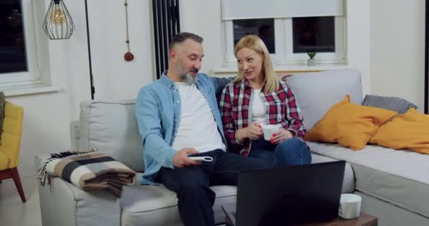 Front view of lovely married adult joyful couple which sitting together on soft danet in living-room and talking ενώ παρακολουθείτε ψυχαγωγικό πρόγραμμα online στο laptop — Αρχείο Βίντεο