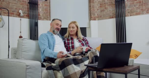 Retrato de casal adulto despreocupado amoroso agradável que relaxa no sofá macio coberto com cobertor e desfrutando de filme interessante no computador — Vídeo de Stock