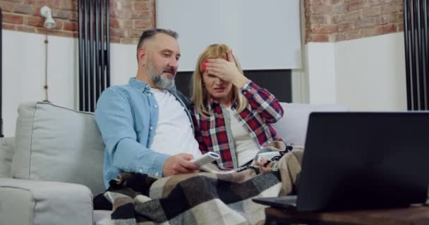 Pareja adulta positiva sin preocupaciones de buen aspecto descansando en un sofá suave en la sala de estar, revisionando la película excitada en el portátil y la mujer cerró los ojos de una escena terrible en la pantalla — Vídeos de Stock