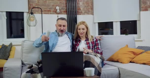 Charmant marié heureux couple joyeux assis sur le canapé à la maison et se réjouissant de balle marqué pendant le match de regarder sur ordinateur pendant la soirée en famille — Video