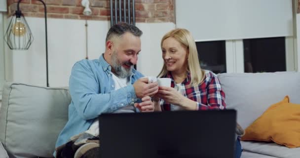 Hermosa pareja adulta feliz amoroso sentado en casa por la noche y tazas de té tintineo durante ver emocionante película en la computadora, concepto de ocio familiar — Vídeos de Stock