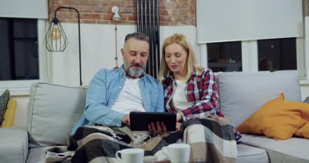 Retrato de bien parecido satisfecho sonriendo feliz pareja de 40-50 que se sientan juntos en el sofá suave en casa y el uso de la tableta PC — Vídeos de Stock