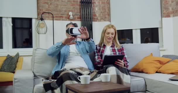 Vue de face du heureux couple marié adulte satisfait qui repose sur un canapé doux dans le salon et en utilisant un casque de réalité virtuelle et un appareil tablette — Video
