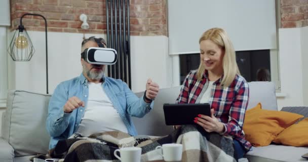 Dobře vypadající spokojený vousatý 50-letý muž ve speciálních 3D brýlích, jako by za volantem, zatímco jeho roztomilá žena sedí vedle něj na pohodlné pohovce a vybírá hru pro něj na tablet PC — Stock video