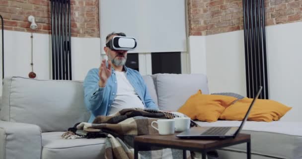Knappe zelfverzekerde serieuze man met baard van 50 jaar in virtual reality headset zittend op een zachte bank thuis en werkend op een denkbeeldig scherm — Stockvideo