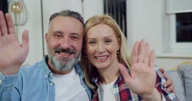 Güzel dekore edilmiş bir dairede kamera önünde el sallayan iyi huylu, mutlu, yetişkin çiftlere yakın durun.