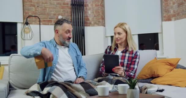 Belle souriante femme heureuse travaillant sur tablette pc tandis que son mari barbu énigmatique donnant sa boîte-cadeau, puis l'embrasser avec amour, concept de famille — Video