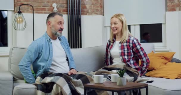 Vue de face d'attrayant sourire heureux couple adulte qui s'assoient ensemble sur le canapé dans son propre appartement, se donnant mutuellement des boîtes-cadeaux et s'embrassant — Video