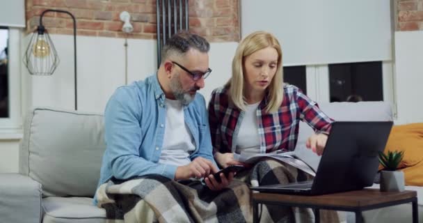 Jolie concentré couple qualifié occupé travaillant ensemble sur ordinateur portable et vérifier les données à l'écran et le rapport, assis sur le canapé à la maison — Video