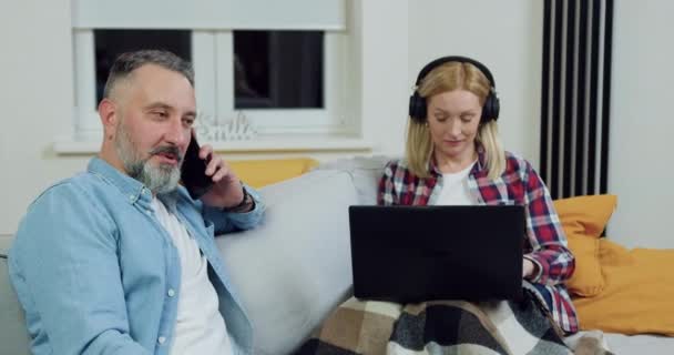 Espléndida mujer adulta sin preocupaciones amorosas en los auriculares que trabajan en el ordenador portátil en la sala de estar y mirando a su marido barbudo experimentado que habla en el móvil — Vídeos de Stock