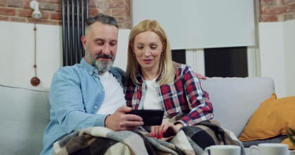 Όμορφος, χαρούμενος, χαρούμενος, ενήλικος γενειοφόρος άντρας και γυναίκα που αναπαύονται στον καναπέ με την κουβέρτα στα πόδια τους και επανεξετάζουν τις προσωπικές τους φωτογραφίες στο κινητό — Αρχείο Βίντεο