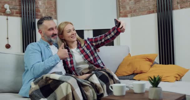Flot smilende glad voksen par sidder på sofaen og gør selfie på smartphone, når du bruger fælles fritid derhjemme – Stock-video