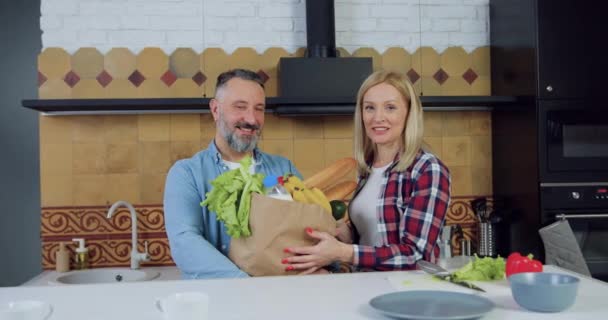 Atractiva sonrisa feliz pareja adulta moderna sosteniendo paquete de alimentos cerca de la mesa y mirando a la cámara en la cocina contemporánea, concepto familiar — Vídeo de stock