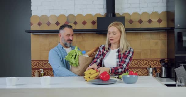 Good-looking loving 45-aged bearded man carrying from market big package with food and his pretty satisfied wife unpacking this package in well-decorated cuisine — Stock Video