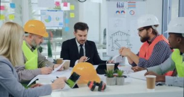 Yakışıklı, sakallı baş şef, kurul odasındaki inşaat planının detaylarını çok ırklı astlarıyla ya da mimarlarıyla tartışıyor.