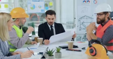 İnşaat şirketinin başarılı sakallı yöneticisi özel üniformalı ve miğferli çok ırklı mühendisleriyle planlarda tasvir edilen mimari projesinin detaylarını tartışıyor.