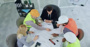 Erkek ve kadın yöneticileriyle toplantı odasındaki proje üzerinde beyin fırtınası yapan yelekli ve kasklı mühendislerden oluşan kendine güveni yüksek profesyonel bir ekip.