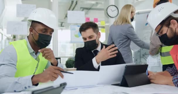 Trabajo durante la pandemia del coronavirus donde los ingenieros multiétnicos confiados y bien parecidos y su director principal o director general en máscaras protectoras hacen una lluvia de ideas sobre el proyecto de construcción durante la reunión — Vídeo de stock