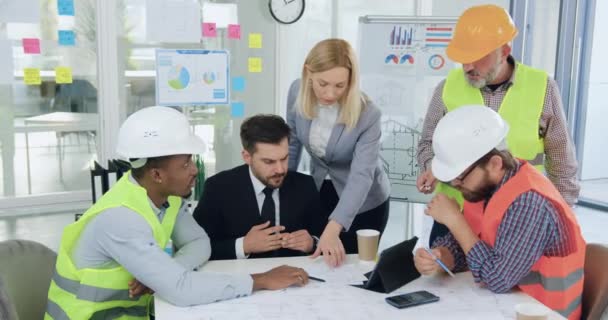 Porträt eines gut aussehenden, hochqualifizierten, zielstrebigen Mischlingsteams aus Bauarbeitern in speziellen Uniformen und den wichtigsten männlichen und weiblichen Managern, die gemeinsam über ein gemeinsames Bauprojekt in — Stockvideo