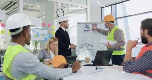 Byggbranschen koncept där snygg skäggig koncentrerad chef diskuterar med äldre entreprenör detaljerna i byggprojekt nära whiteboard medan en annan medlem av laget sitter på — Stockvideo